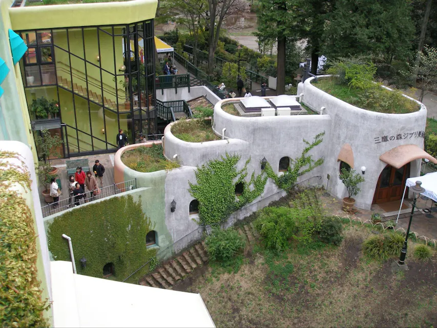 Aerial view of the Studio Ghibli Museum in Mitaka, Japan. The architecture features organic, curved buildings with a green roof and ivy-covered walls. Visitors are walking around the museum's exterior, and a garden area is visible in the foreground.