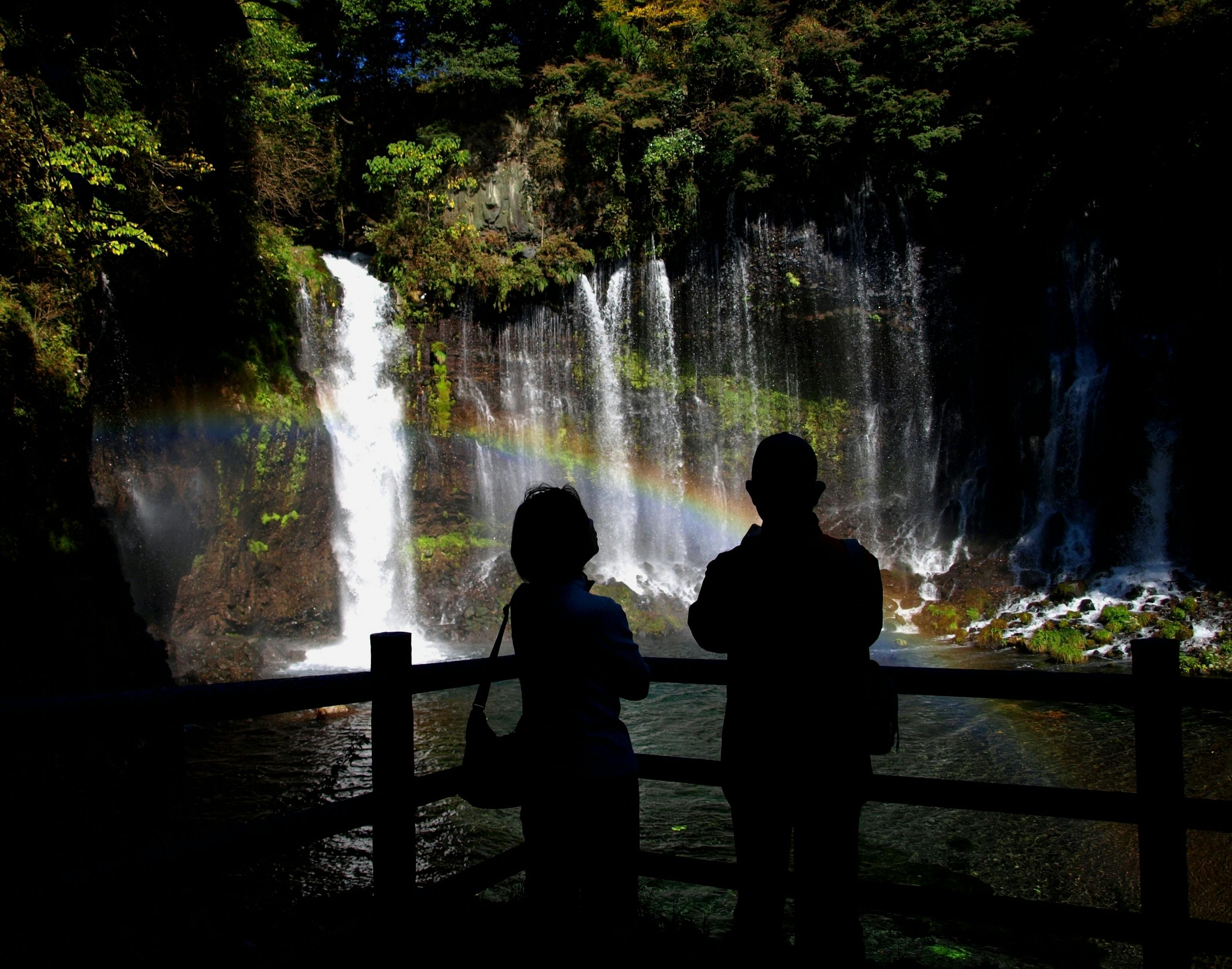 Two people stand in silhouette by a wooden fence, admiring a waterfall and a rainbow forming above the water, surrounded by trees and lush greenery.
