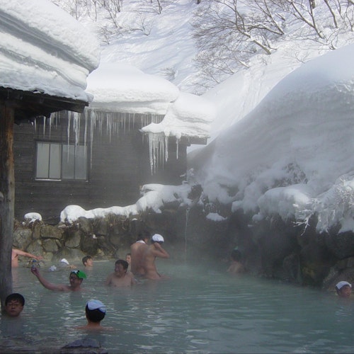 Japan Bathing Culture People enjoying a hot spring surrounded by snow-covered rocks and buildings. Steam rises from the water, and icicles hang from the roof of a nearby structure. Some are wearing towels on their heads, adding to the relaxing atmosphere.