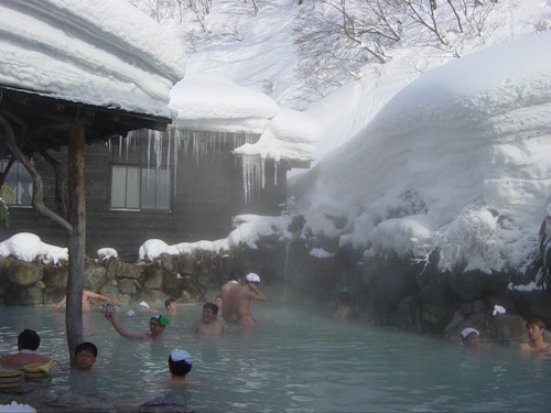 People enjoying a hot spring surrounded by snow-covered rocks and buildings. Steam rises from the water, and icicles hang from the roof of a nearby structure. Some are wearing towels on their heads, adding to the relaxing atmosphere.
