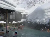 People enjoying a hot spring surrounded by snow-covered rocks and buildings. Steam rises from the water, and icicles hang from the roof of a nearby structure. Some are wearing towels on their heads, adding to the relaxing atmosphere.