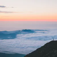 Sea of Clouds A lone hiker stands on a rocky slope overlooking vast, cloud-covered mountains during a beautiful sunrise or sunset. The sky is painted in soft hues of pink, orange, and blue.