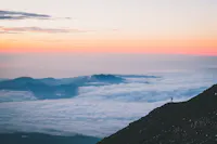 A lone hiker stands on a rocky slope overlooking vast, cloud-covered mountains during a beautiful sunrise or sunset. The sky is painted in soft hues of pink, orange, and blue.