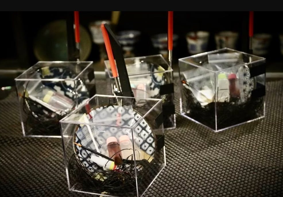 Four clear hexagonal containers hold black shredded material, a test tube with red liquid, and a thin brush with a red handle. Cups with various patterns are blurred in the background.