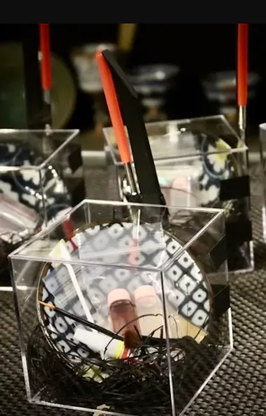 Kintsugi Four clear hexagonal containers hold black shredded material, a test tube with red liquid, and a thin brush with a red handle. Cups with various patterns are blurred in the background.