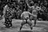 A black and white photo of a sumo wrestling match in an arena. Two sumo wrestlers face each other while a referee stands to the side. The audience watches intently, with many taking pictures and filming the event. The atmosphere is intense and focused.