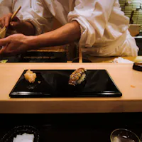 Sukiyabashi Jiro A close-up of a sushi bar with chefs in the background preparing sushi. In the foreground, there is a black rectangular plate with two pieces of sushi. The atmosphere is warm and inviting with partial views of kitchen utensils and ingredients.