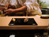 A close-up of a sushi bar with chefs in the background preparing sushi. In the foreground, there is a black rectangular plate with two pieces of sushi. The atmosphere is warm and inviting with partial views of kitchen utensils and ingredients.