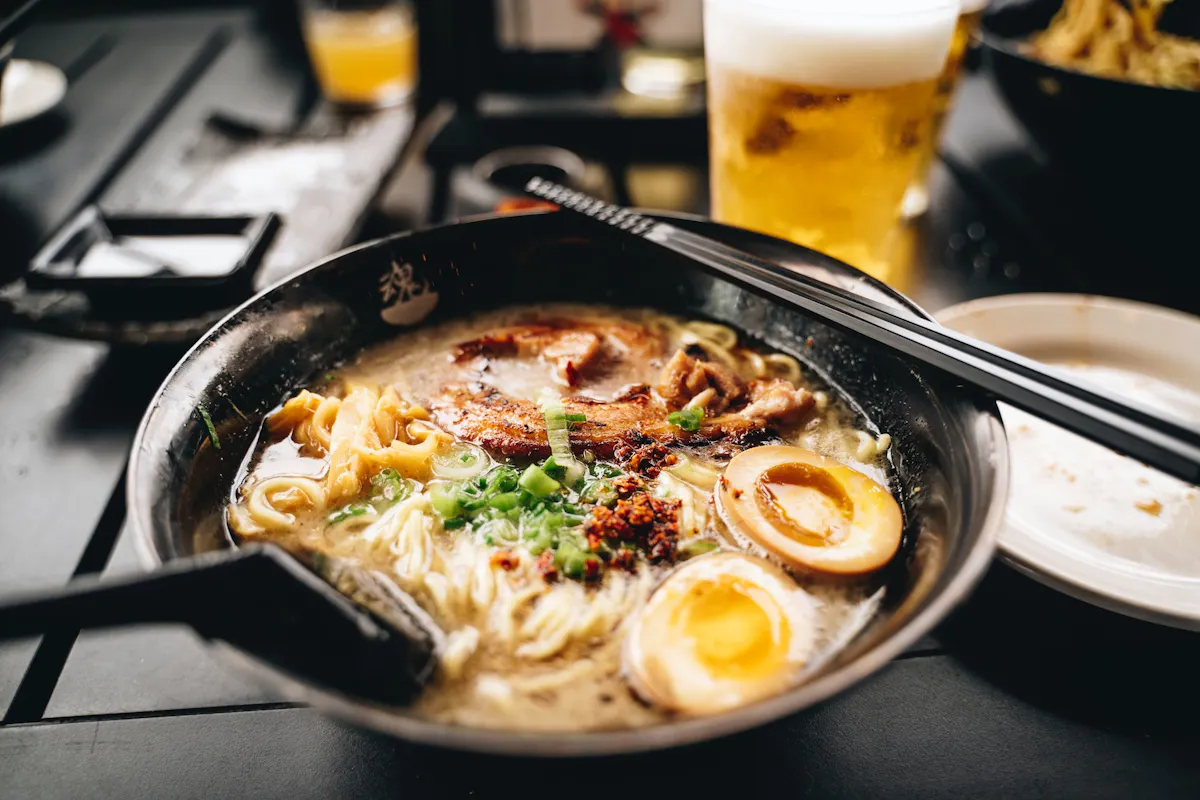 A bowl of ramen with rich broth, sliced meat, soft-boiled egg halves, chopped green onions, and chili flakes. Black chopsticks rest on the bowl, and a blurred glass of beer is in the background on a dark table.