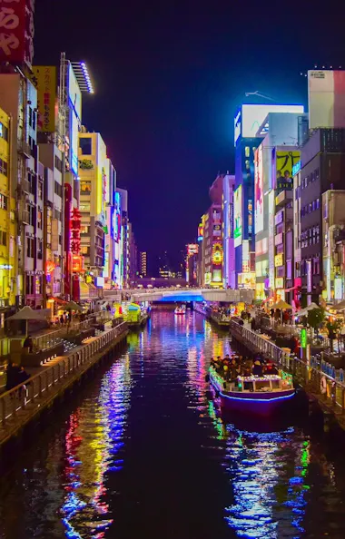 Dotonbori A vibrant cityscape at night shows a canal lined with colorful neon signs, busy sidewalks, and boats on the water, likely in a bustling entertainment district such as Dotonbori in Osaka, Japan.