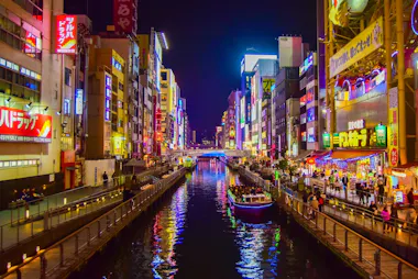 A vibrant cityscape at night shows a canal lined with colorful neon signs, busy sidewalks, and boats on the water, likely in a bustling entertainment district such as Dotonbori in Osaka, Japan.