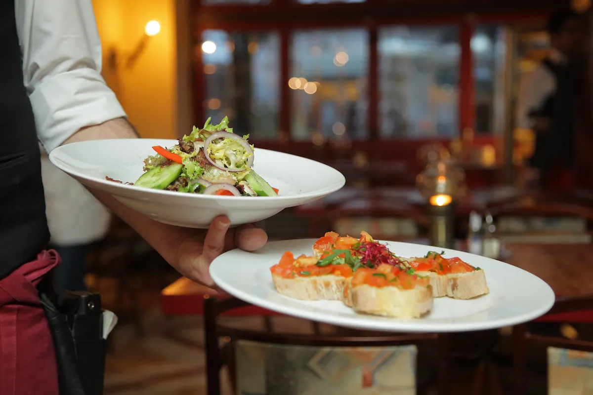 A server in a restaurant carries two plates: one with a salad comprising lettuce, cucumbers, red onions, and tomatoes, and another with bruschetta topped with chopped tomatoes and herbs. The background features a dimly lit, warmly decorated dining area.