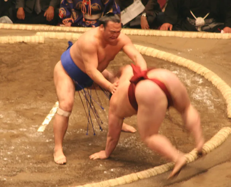 Sumo Wrestling 두 명의 스모 레슬러가 원형 흙 링에서 경기를 합니다. 왼쪽에 있는 레슬러는 파란색 마와시를 입고 붉은색 마와시를 입은 레슬러를 눌러 다치게 하고 있으며, 그 레슬러는 몸을 구부리고 균형을 잃어가는 모습입니다. 배경에는 관중들이 보입니다.