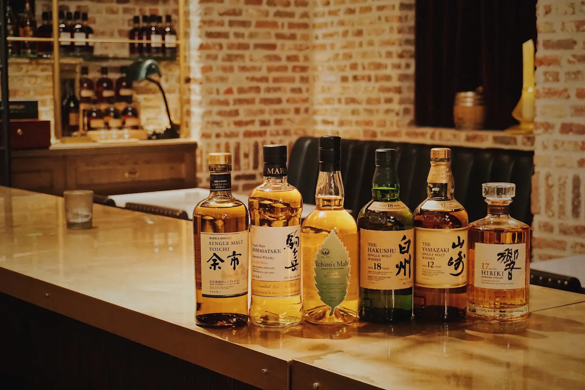 A selection of six whiskey bottles displayed on a polished wooden counter against a backdrop of brick walls and shelves filled with various bottles. The bottles have distinct labels with some featuring English and Japanese text. The setting appears cozy and dimly lit.