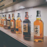 Japanese Whisky A row of various whisky bottles displayed on a backlit shelf. The bottles have labels with Japanese writing and are arranged in a line, each slightly differing in color. The shelf is part of a store or tasting room with a modern and organized layout.