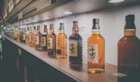 A row of various whisky bottles displayed on a backlit shelf. The bottles have labels with Japanese writing and are arranged in a line, each slightly differing in color. The shelf is part of a store or tasting room with a modern and organized layout.