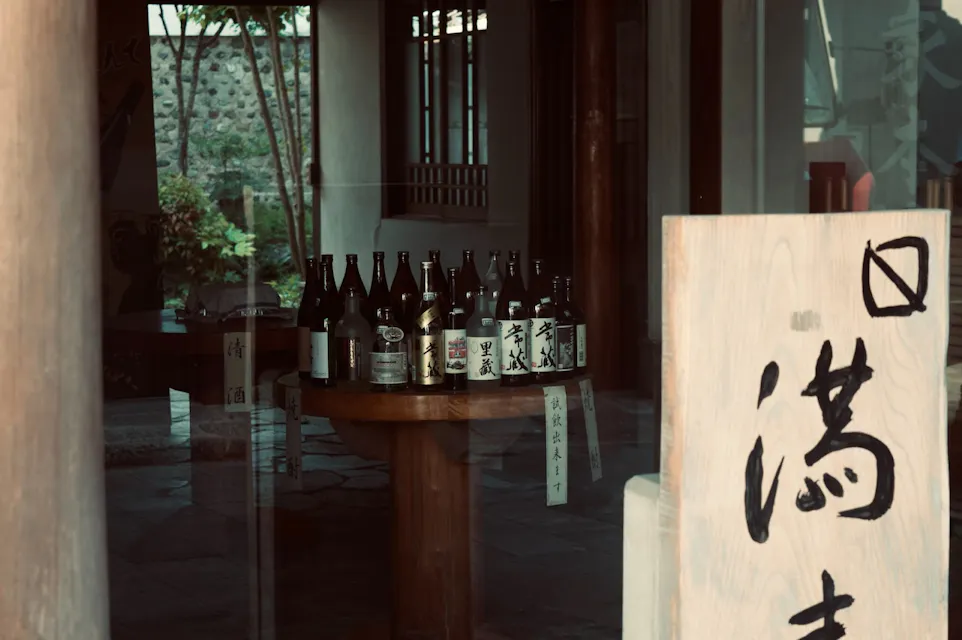 Japanese Whisky A wooden table in a dimly lit room holding multiple bottles, possibly of sake, is seen through a window. There is traditional Japanese calligraphy on a large vertical wooden sign in the foreground, partially obscuring the view.