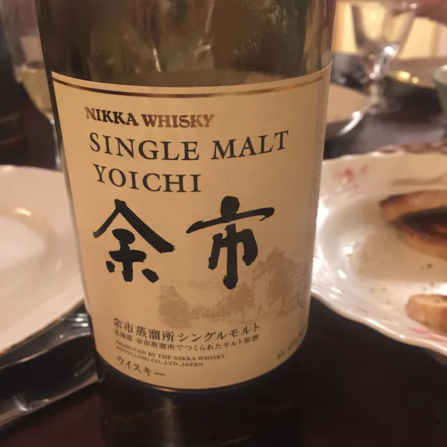 Japanese Single Malt Whisky A bottle of Nikka Whisky Single Malt Yoichi is placed on a table. The brown glass bottle features a label with Japanese characters and English text. Surrounding the bottle are plates with remnants of food and glasses, indicating a meal or gathering setting.
