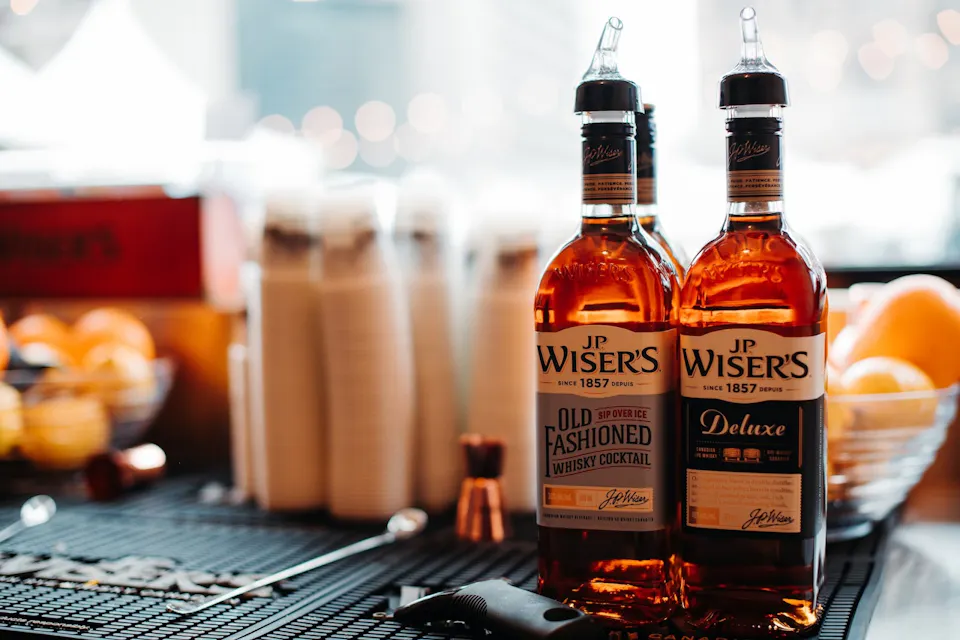 Japanese Whisky Two bottles of JP Wiser's whisky, labeled "Old Fashioned Whisky Cocktail" and "Deluxe," stand on a bar counter. Behind them are plastic cups, and to the right, a bowl of oranges. The background is softly blurred, highlighting the bottles in the foreground.