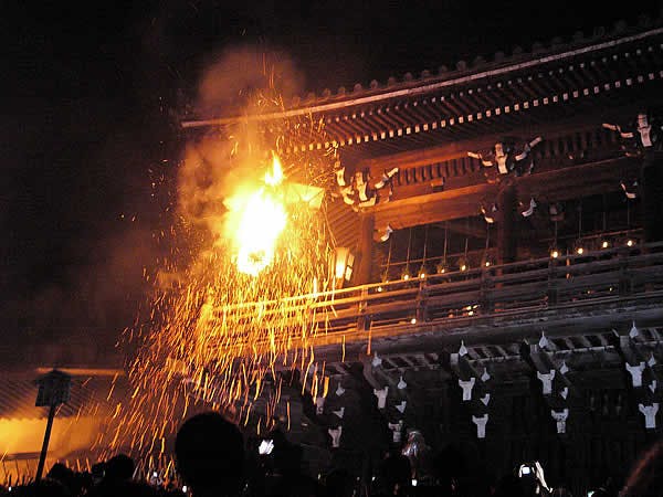A large fire torch emits bright sparks from a wooden balcony of a traditional Japanese temple at night, with people watching the event below.