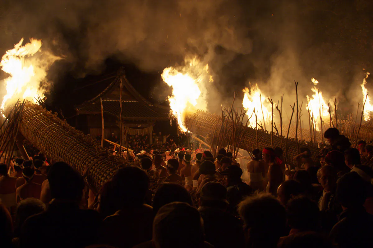 Oniyo Fire Festival A large crowd gathers at night for a fire festival, with massive torches and bonfires illuminating the scene. Participants hold smaller torches and wear traditional attire, while onlookers gather around to watch the event. A shrine is visible in the background.