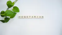 A green leafy plant on the left side extending across a white background. The word "VEGETARIAN" is spelled out in black letters on individual white tile pieces, positioned horizontally in the center of the image.