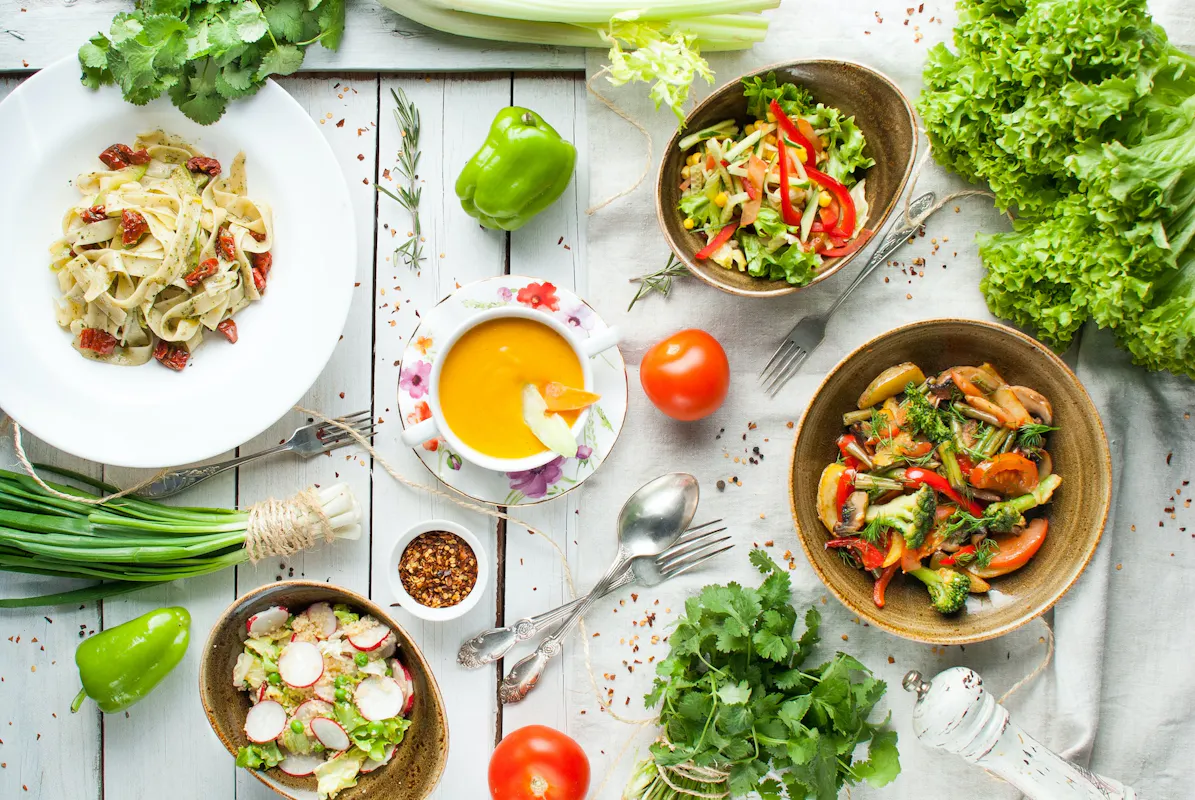 A variety of dishes on a rustic white wooden table includes pasta with sundried tomatoes, vegetable soup, mixed salad, and a stir-fry with broccoli and bell peppers. Surrounding ingredients include green onions, cilantro, celery, bell pepper, lettuce, and tomatoes.