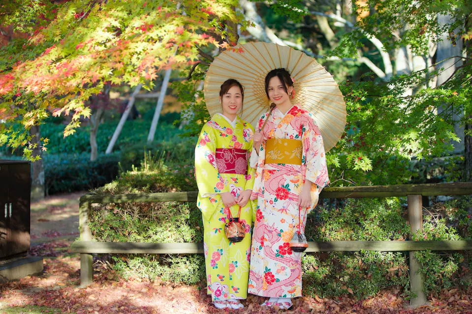 两个穿着色彩斑斓和服的女人站在秋天树叶的花园里。一位女性手持阳伞，她们俩对着镜头微笑。花园中有生机勃勃的绿叶和红叶，背景是一道木栅栏。