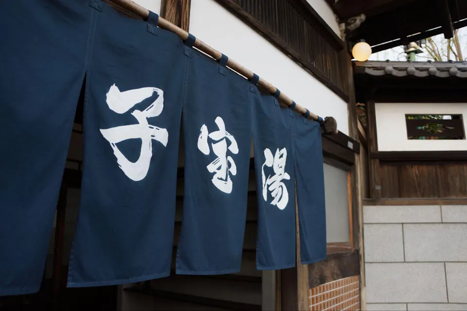 一幅传统日本玄关帷幕（称为暖帘）的照片，悬挂在一座木质建筑前。帷幕深蓝色，上面画有白色日文汉字，表示该场所的名称或功能。