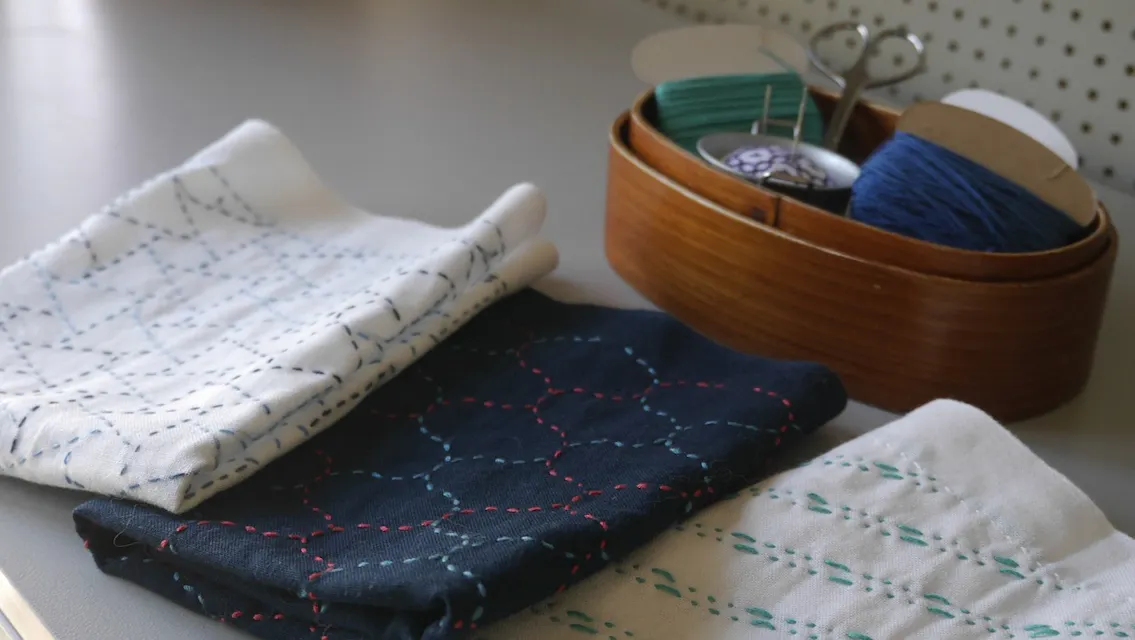 一组折叠面料的特写，展示了蓝色、红色和绿色线的Sashiko刺绣图案。背景中是一个装有线轴、剪刀和针的木容器， resting 在灰色表面上。