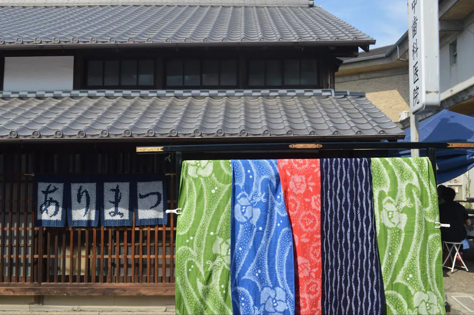 背景中有一座传统的日本建筑，屋顶铺有瓦片。在前面，各种图案和设计的彩色布料片悬挂着。在左侧，展示了一条部分被阴影遮挡的带有日文字母的布条。