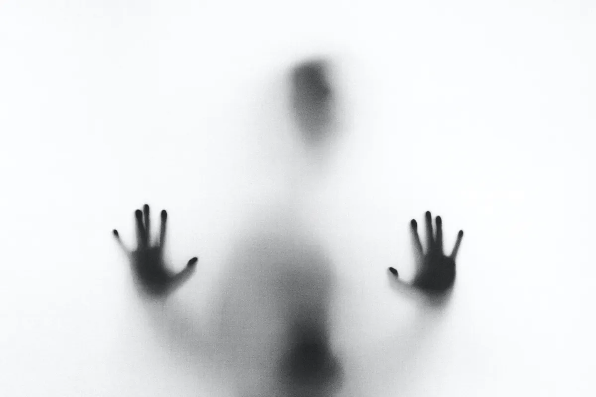 Paranormal Image A blurry silhouette of a person with their hands pressed against a frosted or opaque surface. The image captures an ambiguous and mysterious figure, with the details of the face and body obscured.
