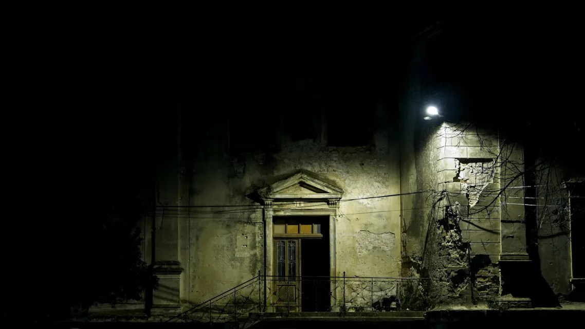 An old, abandoned building is partially illuminated by a single street lamp at night. The facade shows signs of decay, with cracked walls and peeling paint. The entrance features a weathered wooden door and some stairs leading up to it.