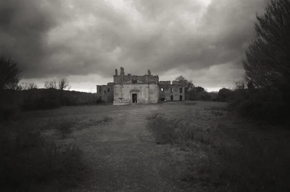 A black and white photo of a dilapidated, abandoned building under a cloudy, ominous sky. A dirt path leads up to the building, which appears to be missing a roof and has crumbling walls. Leafless trees surround the structure, adding to the eerie atmosphere.