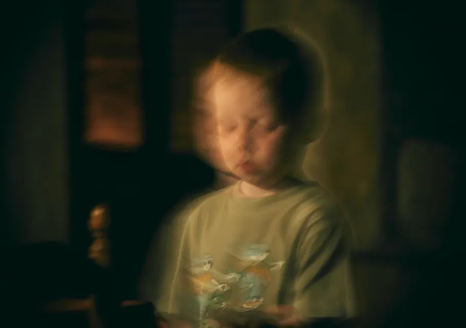 A blurred, ethereal image shows a young child with red hair standing indoors. The child's face and surroundings are indistinct, giving a ghostly effect. The child is wearing a light-colored shirt with a cartoon graphic. Dark, muted background.