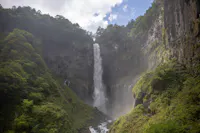 A tall, narrow waterfall cascades down a rocky cliff covered with greenery into a misty basin below. The surrounding cliffs are lush with vegetation, and the sky above is partly cloudy, allowing sunlight to illuminate parts of the scene.