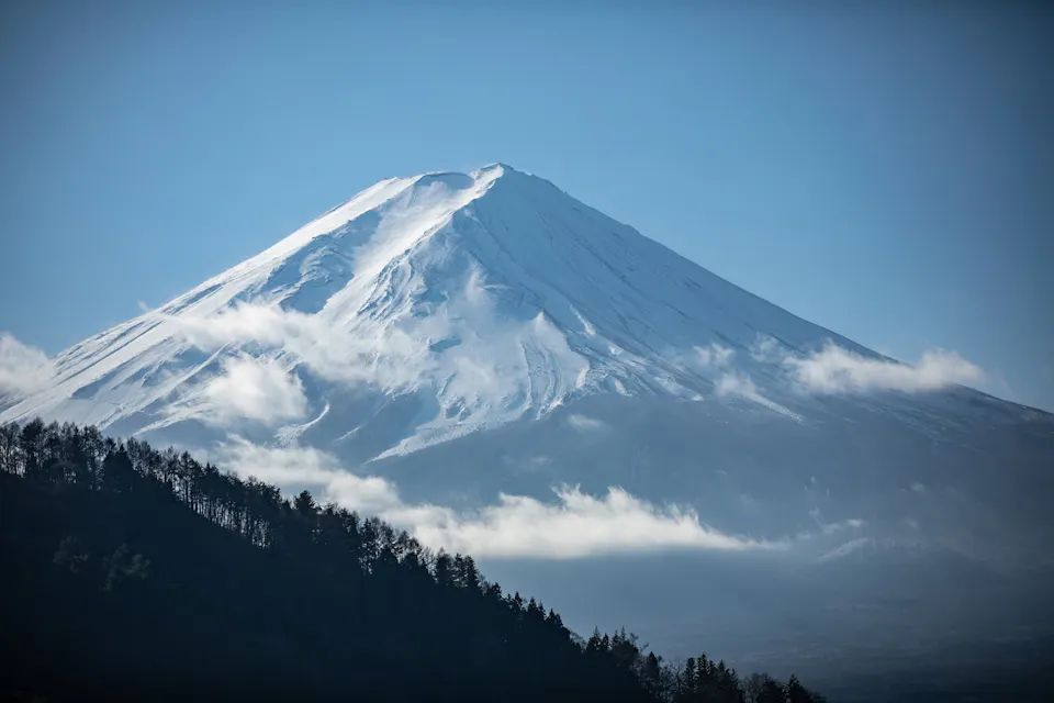 雪に覆われた富士山が晴れた青空を背景にそびえ立っています。山の頂は新しい雪でキラキラ光り、軽い雲が部分的に山を包み込んでいます。前景には、暗い森に覆われた ridge が明るい雪と対照をなしています。