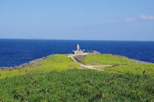A scenic view of Cape Manzamo in Okinawa, Japan, with lush green grass in the foreground, a clear blue sky above, and the deep blue ocean in the background. A pathway leads to a small monument on a rocky outcrop, where several people are gathered.