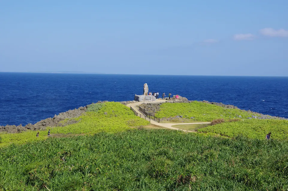 A scenic view of Cape Manzamo in Okinawa, Japan, with lush green grass in the foreground, a clear blue sky above, and the deep blue ocean in the background. A pathway leads to a small monument on a rocky outcrop, where several people are gathered.