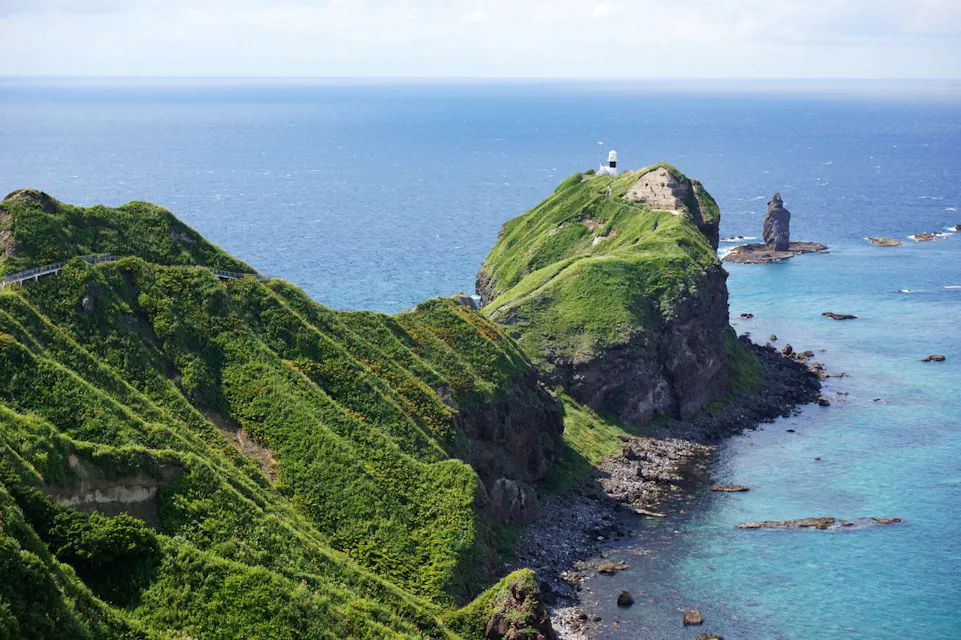 A lush, green peninsula with steep slopes extends into a blue ocean under a partly cloudy sky. A small white lighthouse sits atop the cliff at the peninsula's end, while a solitary rock formation stands in the water nearby. The coastline is peppered with rocky outcrops.