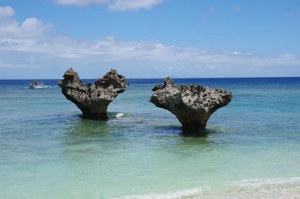 Two uniquely shaped rock formations stand in the shallow, clear turquoise water along a coastline under a vibrant blue sky with scattered clouds. In the distance, a small boat is visible on the left side, adding a sense of scale to the serene scene.