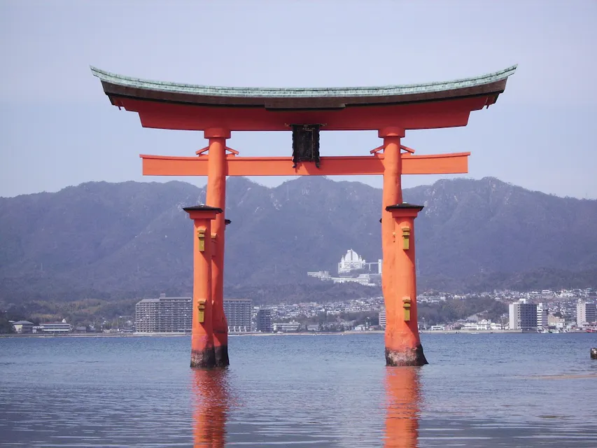 A large, traditional Japanese torii gate stands in the water, with a mountainous landscape and buildings visible in the background. The bright orange hue of the gate contrasts with the calm blue water and the clear sky.