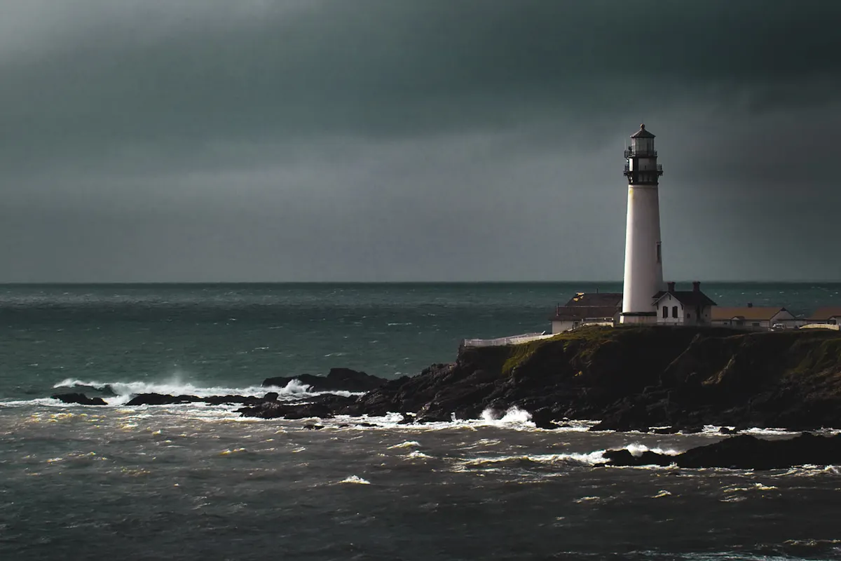 Lighthouse A solitary lighthouse stands on a rocky cliff overlooking a turbulent sea under a cloudy, dark sky. Waves crash against the rocks, and a few buildings are visible near the base of the lighthouse. The atmosphere is moody and dramatic.