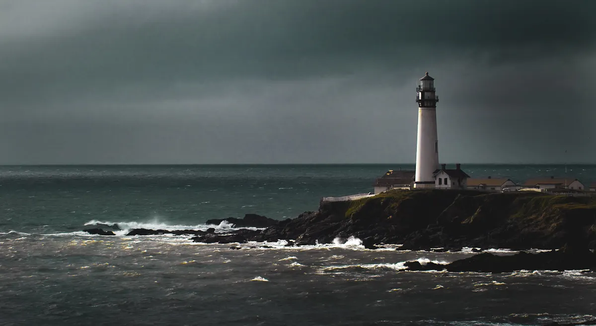 A solitary lighthouse stands on a rocky cliff overlooking a turbulent sea under a cloudy, dark sky. Waves crash against the rocks, and a few buildings are visible near the base of the lighthouse. The atmosphere is moody and dramatic.