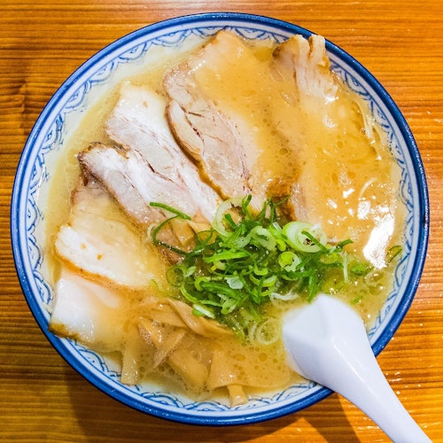 Ramen Paradise A bowl of ramen with slices of chashu pork, sliced green onions, and a white ceramic spoon resting on the edge. The ramen has a rich, creamy broth and is served in a patterned blue and white bowl, placed on a wooden surface.