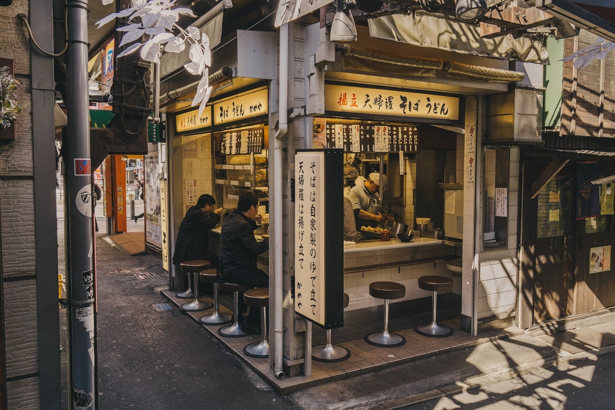 Japanese Street Food A small Japanese noodle shop with seating at a counter facing the street, partially shaded by overhanging structures. Several patrons sit at the counter eating, while the interior is brightly lit. The exterior features signage and traditional decor.