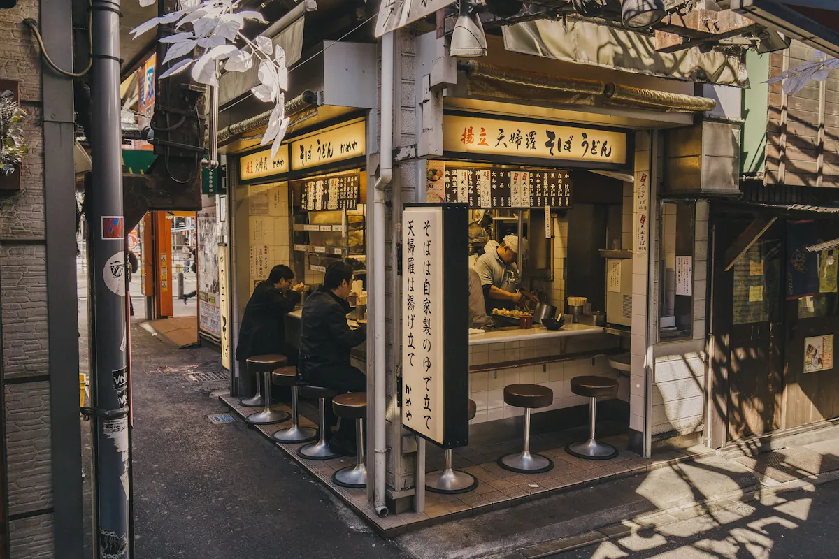 Japanese Street Food A small Japanese noodle shop with seating at a counter facing the street, partially shaded by overhanging structures. Several patrons sit at the counter eating, while the interior is brightly lit. The exterior features signage and traditional decor.