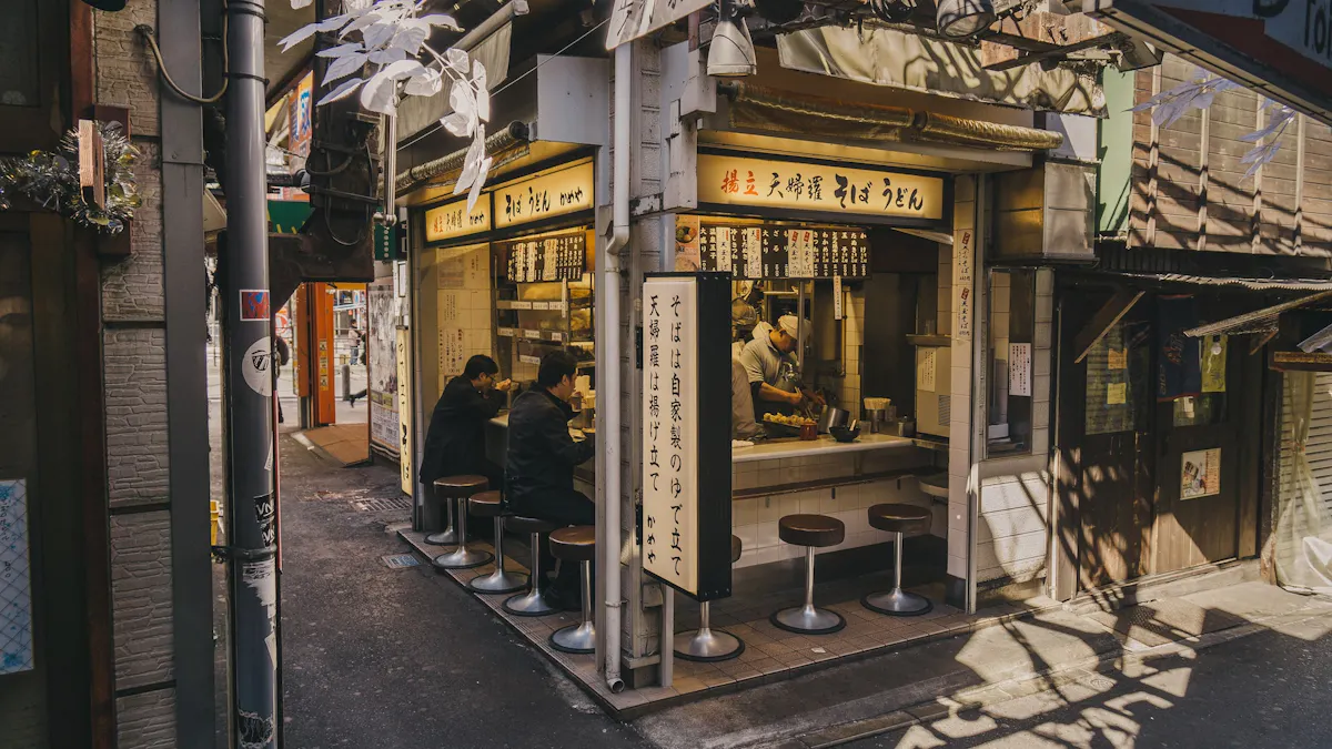 A small Japanese noodle shop with seating at a counter facing the street, partially shaded by overhanging structures. Several patrons sit at the counter eating, while the interior is brightly lit. The exterior features signage and traditional decor.