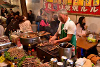 A street vendor in a green apron is grilling skewered meat at a busy night market. The stall is crowded with customers seated at tables enjoying their meals. Various condiments, cans, and cooking utensils are scattered around, with a vibrant banner displayed in the background.