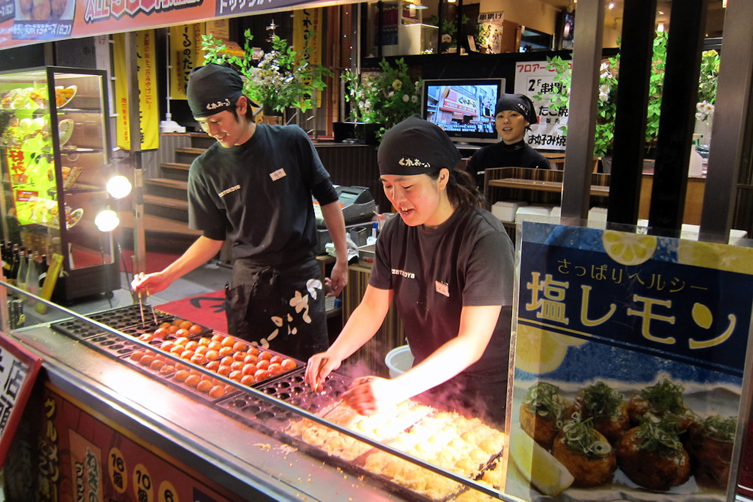 Two people in black uniforms and hats are cooking takoyaki (octopus balls) at a street food stall in Japan. The scene is vibrant, with the grill and food display in the foreground. A third person in the background appears to be smiling.